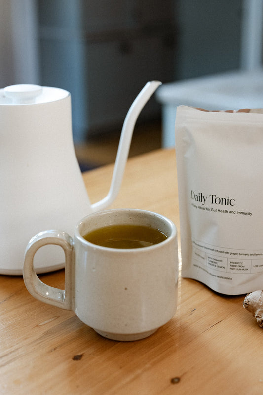 Best Bones Broth Daily Tonic in a ceramic mug beside a gooseneck kettle and fresh ginger on a wooden bench — a daily ritual for gut health and immunity